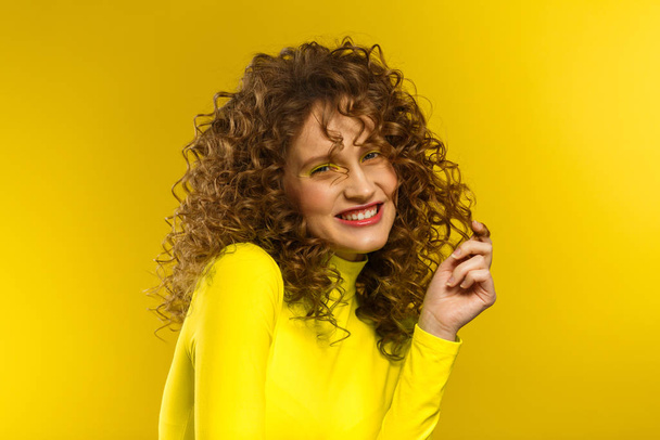 Portrait of young beautiful smiling women with curly hair. Happy girl with wavy hairstyle and bright make up isolated over yellow background. Beauty and hair care concept. Yellow lifestyle. Copy space - Fotografie, Obrázek