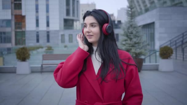 Portrait of young cheerful Caucasian woman dancing in earphones on city street. Elegant girl in red coat listening to music in headphones. Lifestyle, hobby, joy. - Video, Çekim