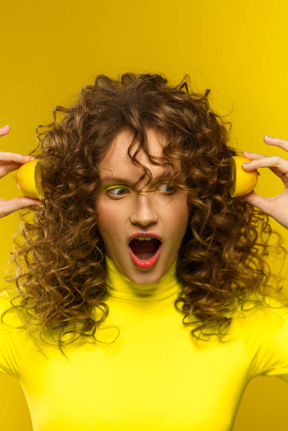 Beautiful curly-haired women with open mouth holding lemon halves near ears, being surprised and shocked isolated over yellow background. Reaction on unexpected news. Copy space. Expressive faces - Fotoğraf, Görsel