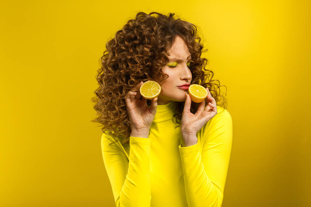 Young beautiful smiling women with curly hair tasting sour lemon halves emotionally. Funny girl with wavy hairstyle and bright make up isolated over yellow background. Copy space. Yellow lifestyle - Zdjęcie, obraz