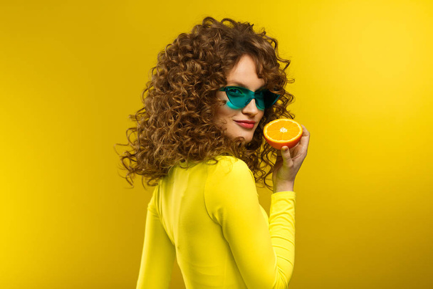 Young beautiful smiling women with curly hair tasting sour lemon halves emotionally. Funny girl with wavy hairstyle and bright make up isolated over yellow background. Copy space. Yellow lifestyle - Photo, image
