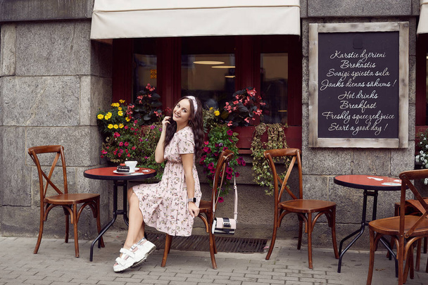Young beautiful bright girl in a summer dress sitting at a European cafe table - Valokuva, kuva