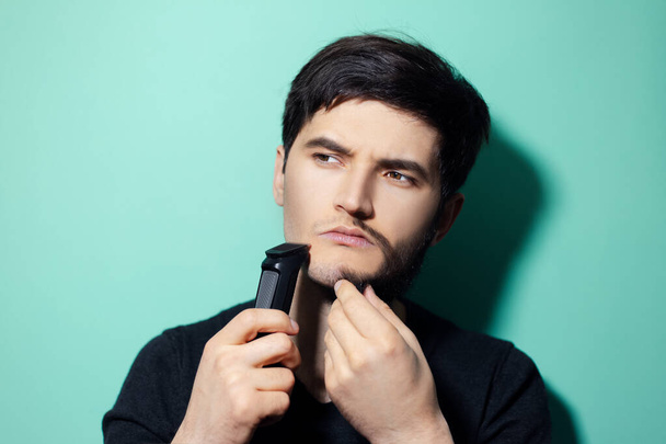 Studio portrait of young man with half shaved face, holding electric shaver trimmer on background of aqua menthe color. - Foto, Imagen