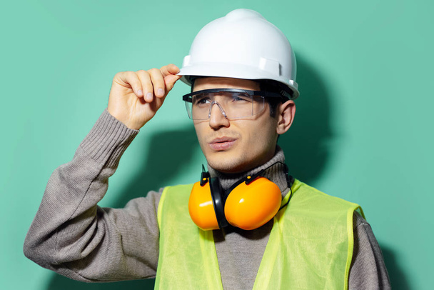 Studio portrét mladého stavitele, inženýra, dotýkající se jeho bezpečnostní přilby pro stavbu, nošení brýlí a sluchátek na pozadí aqua men barvy. - Fotografie, Obrázek