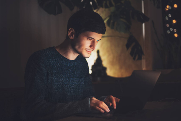 Νυχτερινό πορτρέτο του νεαρού χαρούμενου τύπου που εργάζεται σε φορητό υπολογιστή σε σκοτεινό δωμάτιο με γιρλάντες στο τραπέζι στο σπίτι. - Φωτογραφία, εικόνα