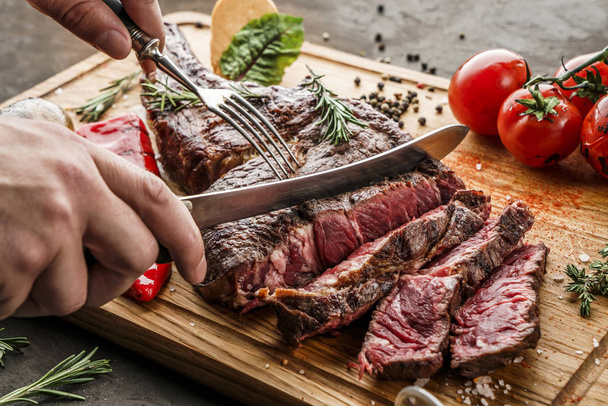 Hands cut grilled tomahawk meat medium rare or rib eye steak on wooden cutting board with grilled vegetables on dark background, close up - Photo, Image