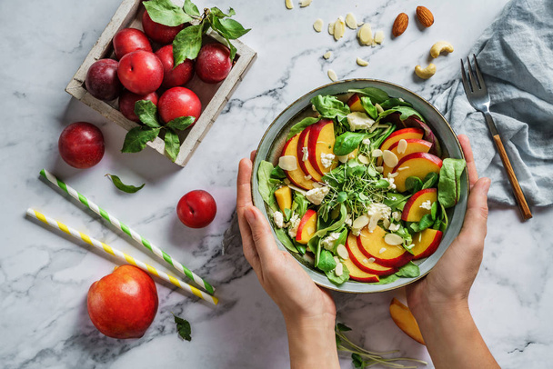 Hands holding fresh summer salad with peach, spinach, micro greens, plums, feta cheese and almonds on light marble background. Healthy food, clean eating, Buddha bowl salad, top view - Fotografie, Obrázek