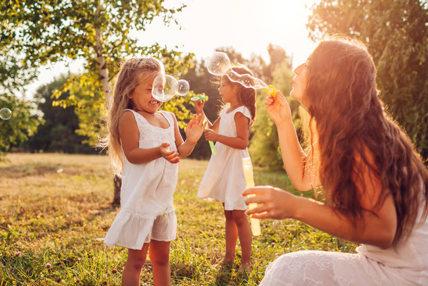 Dia da mãe. Mulher ajuda filhas a soprar bolhas de sabão no parque de verão. Crianças se divertindo jogando ao ar livre
 - Foto, Imagem