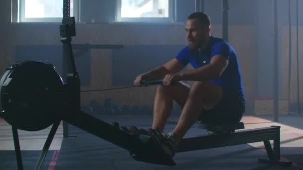 Een sterke man in slow motion trekt aan een touw in een roeisimulator. Cardio-training voor één man in een sfeervolle fitnessruimte. - Video