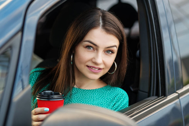 Μια επιτυχημένη γυναίκα κάθεται σε ένα αυτοκίνητο και πίνει ζεστό καφέ. Η έννοια της μεταφοράς, της γυναίκας των επιχειρήσεων και της ασφάλισης. - Φωτογραφία, εικόνα