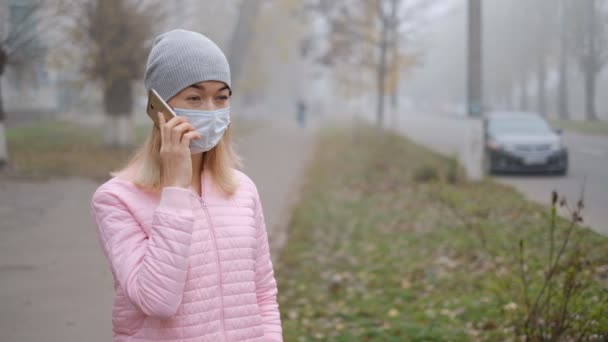 Protection contre le coronavirus. Une jeune femme portant un masque de protection médicale se tient avec un smartphone dans une rue de la ville en Europe. - Séquence, vidéo