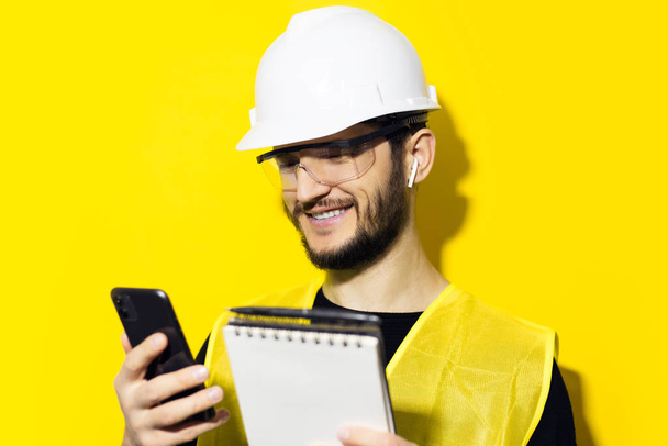 Studio portrét mladého usměvavého muže architekta, stavebního inženýra, v bílé stavební bezpečnostní přilbě, brýlích a bundě. Použití smartphone a bezdrátové sluchátka, izolované na žlutém pozadí. - Fotografie, Obrázek