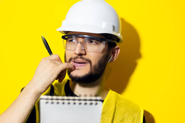 Retrato del estudio de un joven pensante, ingeniero constructor con casco de seguridad y gafas para la construcción sobre fondo amarillo. Sosteniendo un bolígrafo y un cuaderno
. - Foto, Imagen