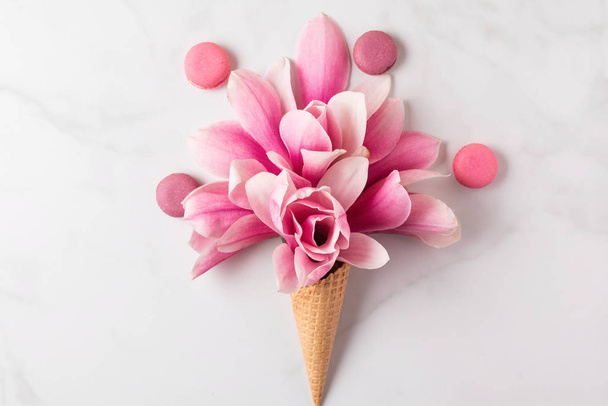 Creative layout made with pink magnolia flowers in waffle cone with macaroons on white marble background. Flat lay - Photo, Image