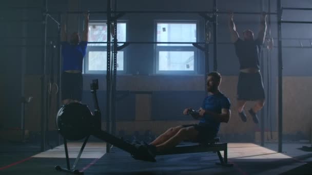 Three men work out together in a fitness room. A man pulls a rowing machine, and two perform pull-UPS on the bar. Cross-training in slow motion. - Footage, Video