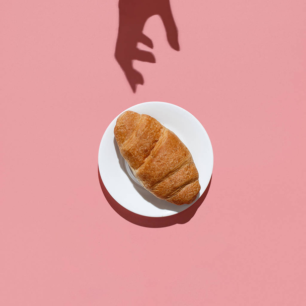 The shadow of the womans hand reaches for a large croissant on a pink background. Directly above flat lay - Фото, зображення