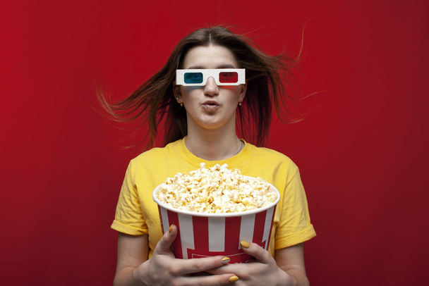 surprised young girl student watching a movie in 3D glasses and eating popcorn on a red background - Photo, Image