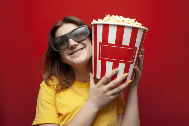 happy young girl the viewer in 3D glasses holds a big box of popcorn and watches a movie on a red color background - Photo, Image