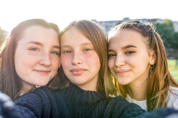 Trzy nastoletnie dziewczyny w wieku 12-14 lat, letnie miasto oglądające filmy telefoniczne, szczęśliwe uśmiechnięte osoby bawiące się radują. Selfie portret z bliska. Zwykłe ubrania, swetry. Emocje pozytywnego relaksu. - Zdjęcie, obraz