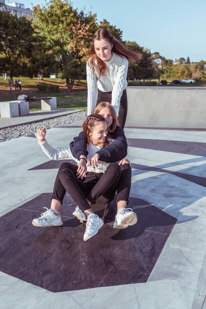 Three teenage girls of schoolgirls 12-14 years old, in summer in city, ride skateboard, happy smiling, having fun rejoicing. Weekend break. Casual clothes, warm sweaters. Emotions positive and delight - Фото, изображение