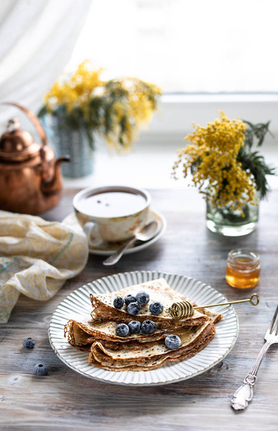 A plate with pancakes with blueberry berries on a wooden table. In the background is a cup of tea and a bouquet of spring flowers by the window. - Photo, Image