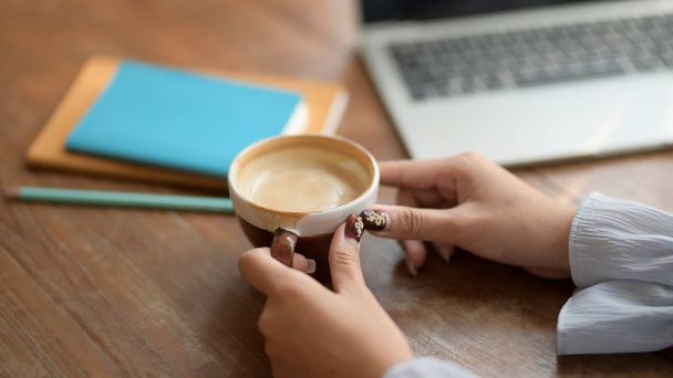 Vista da vicino di una donna che tiene una tazza di caffè in un posto di lavoro confortevole con laptop e cancelleria
 - Foto, immagini
