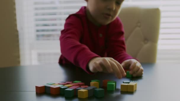 small boy in a red sweater is playing with the colorful wooden blocks on a table in front of a window.Closeup video. - Кадры, видео