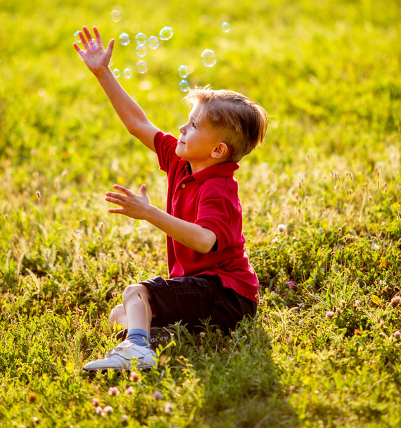 Boy sitting on green grass outdoor playing with soap bubbles - Fotoğraf, Görsel