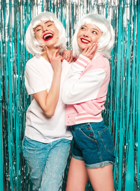 Twee jonge sexy lachende hipster meisjes in witte pruiken en rode lippen.Mooie trendy vrouwen in de zomer kleding.Zorgeloos modellen poseren op blauw zilver glanzende tinsel achtergrond in studio.Positieve vrouwelijke - Foto, afbeelding