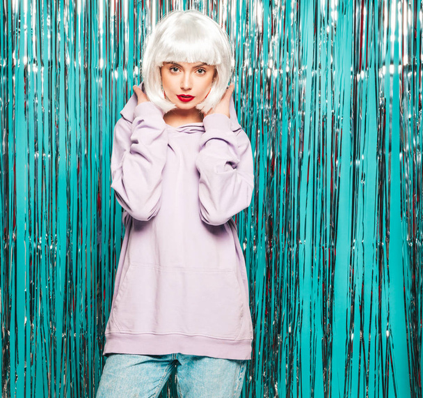 Young sexy smiling hipster girl in white wig and red lips.Beautiful trendy woman in summer clothes.Carefree model posing on blue silver shiny tinsel background in studio.Positive female going crazy - 写真・画像