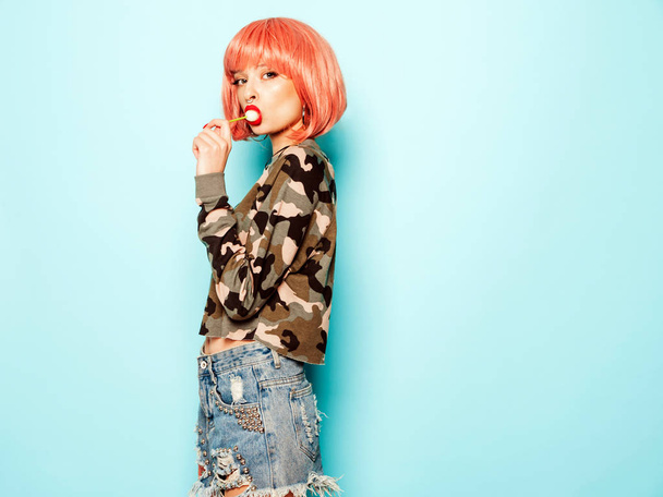 Portrait of young beautiful hipster bad girl in trendy jeans shorts and earring in her nose.Sexy carefree smiling woman in pink wig posing in studio.Positive model licking round sugar candy - Φωτογραφία, εικόνα