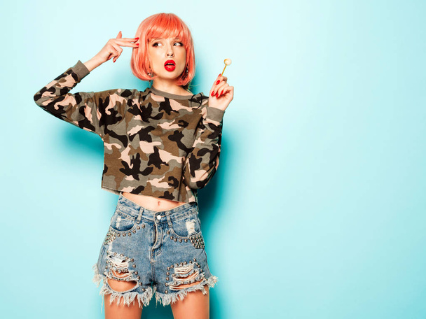 Portrait of young beautiful hipster bad girl in trendy jeans shorts and earring in her nose.Sexy carefree smiling woman in pink wig posing.Positive model licking round sugar candy.Makes gun sign - Фото, зображення