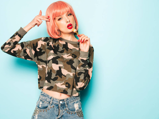 Portrait of young beautiful hipster bad girl in trendy jeans shorts and earring in her nose.Sexy carefree smiling woman in pink wig posing.Positive model licking round sugar candy.Makes gun sign - 写真・画像