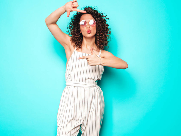 Portrait of beautiful smiling model with afro curls hairstyle dressed in summer hipster clothes.Sexy carefree girl posing near blue wall.Trendy woman makes photo frame by hands - Фото, изображение