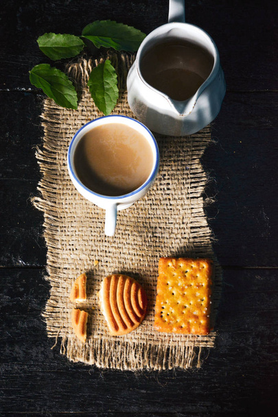 A cup of tea with tasty biscuits, teapot and fresh Leaves on old wooden dark background - Photo, Image