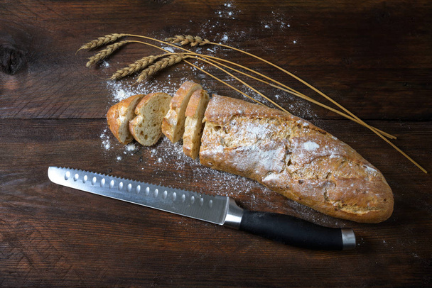 Baguette or French bread half sliced, some wheat ears and a knife on a dark rustic wood, copy space, high angle view from above - Fotoğraf, Görsel