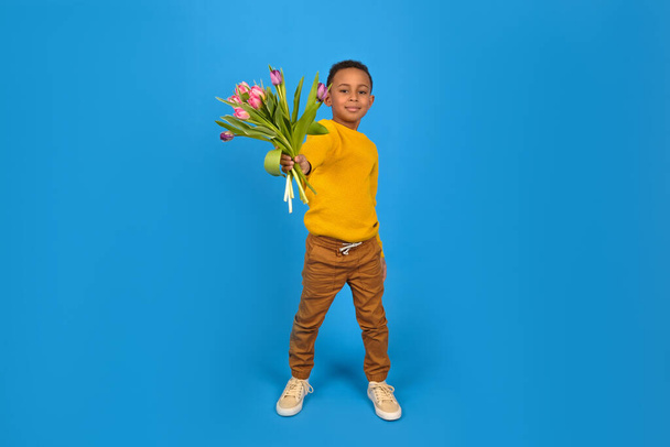 Portrait of a smiling African American boy with a bouquet of tulips for mom spring flowers on a blue background. Concept of greeting mothers Day, fathers day, Valentines day gift for a woman. - Фото, изображение