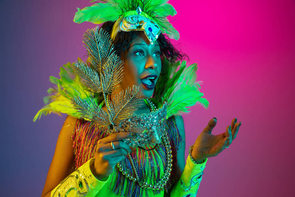 Beautiful young woman in carnival and masquerade costume on gradient studio background in neon light - 写真・画像