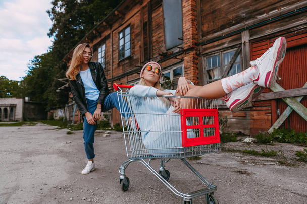 A long haired woman wearing a powder blue dress, pink woollen hat is chilling with her female friend, who is sitting on a shopping trolley, wearing a black leather jacket and blue jeans, in a derelict - Fotó, kép