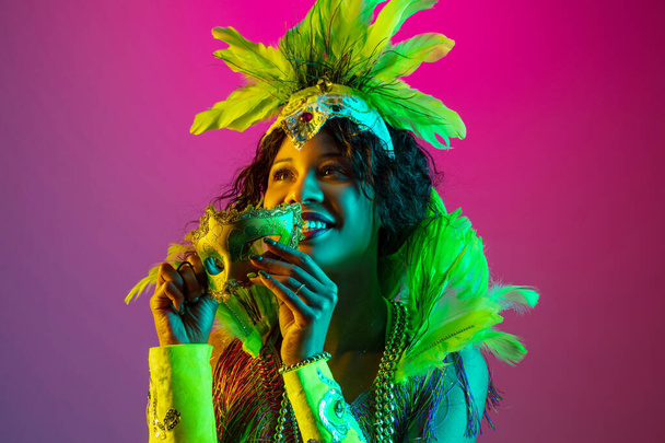 Beautiful young woman in carnival and masquerade costume on gradient studio background in neon light - Fotó, kép