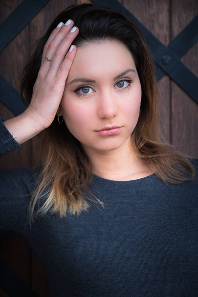 Attractive young brunette woman with large grey blue eyes and long hair looking solemnly at the camera with her hand to her head against a wooden wall - Photo, Image