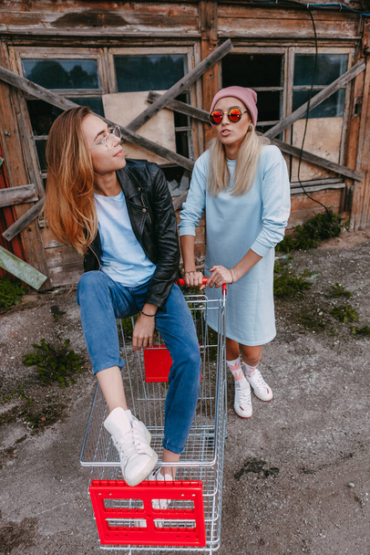 A long haired woman wearing a powder blue dress, pink woollen hat is chilling with her female friend, who is sitting on a shopping trolley, wearing a black leather jacket and blue jeans, in a derelict - Foto, afbeelding