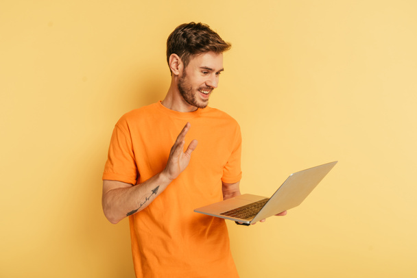 веселий молодий чоловік махає рукою, маючи відеодзвінок на ноутбук на жовтому фоні
 - Фото, зображення