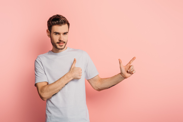 serious young man showing thumb up and gun gesture while looking at camera on pink background - Foto, Bild