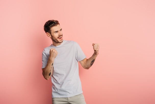excited young man showing winner gesture while looking away on pink background - Photo, Image