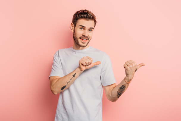 cheerful young man looking at camera while pointing with thumbs on pink background - Photo, Image