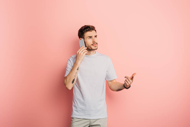 thoughtful young man looking away while talking on smartphone on pink background - Fotoğraf, Görsel