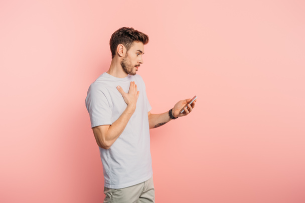 sorprendido joven tocando el pecho mientras sostiene el teléfono inteligente sobre fondo rosa
 - Foto, imagen
