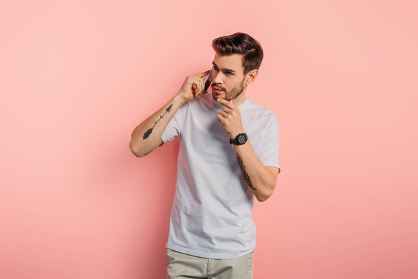 thoughtful young man touching chin while talking on smartphone on pink background - Foto, Bild