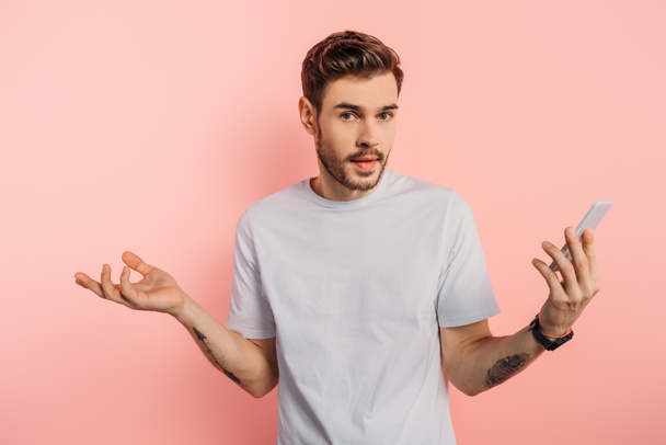 confused young man showing shrug gesture while holding smartphone on pink background - Фото, зображення
